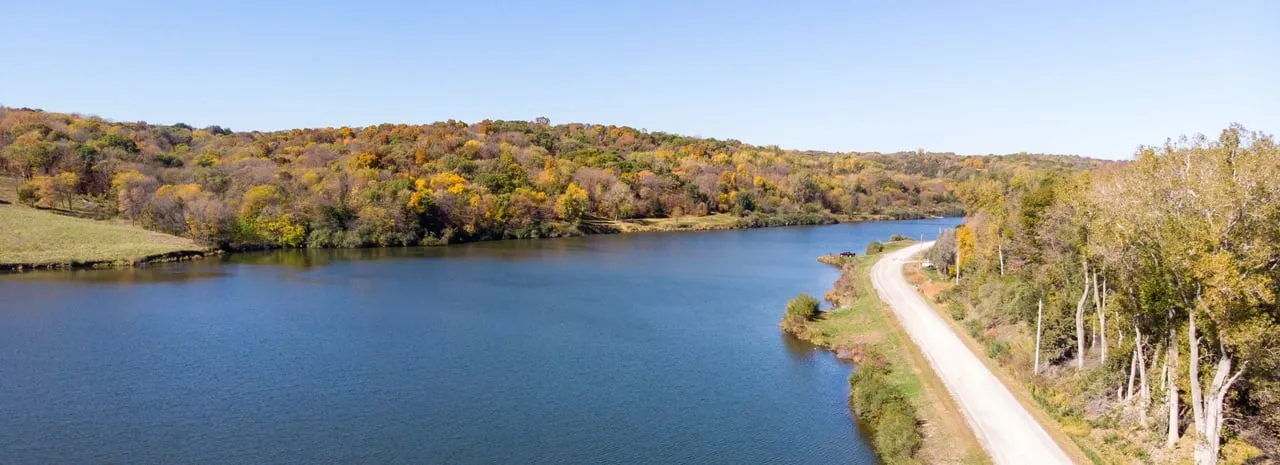 Aerial view of a lake and curving shoreline road, bordered by autumn-colored trees.
