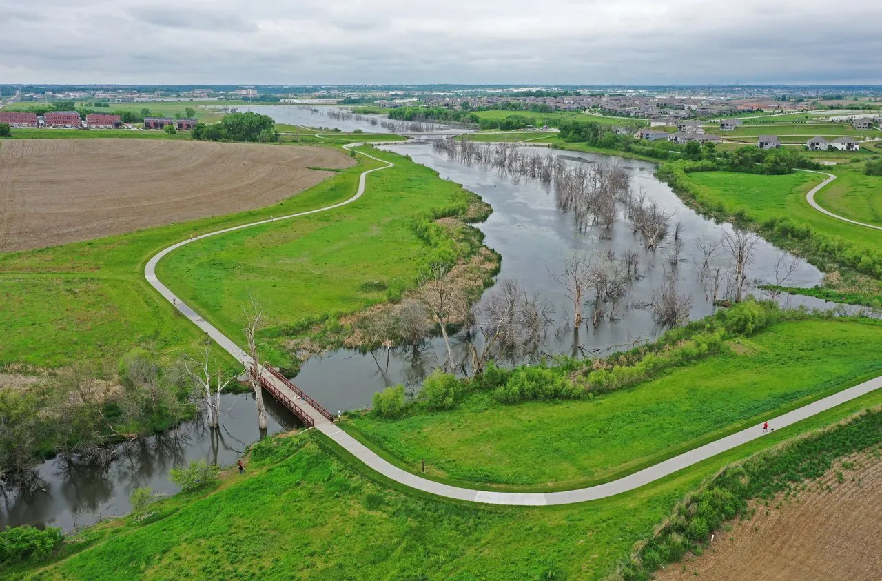 Aerial view of a river winding through green parkland, with trails, a footbridge, and homes beyond.