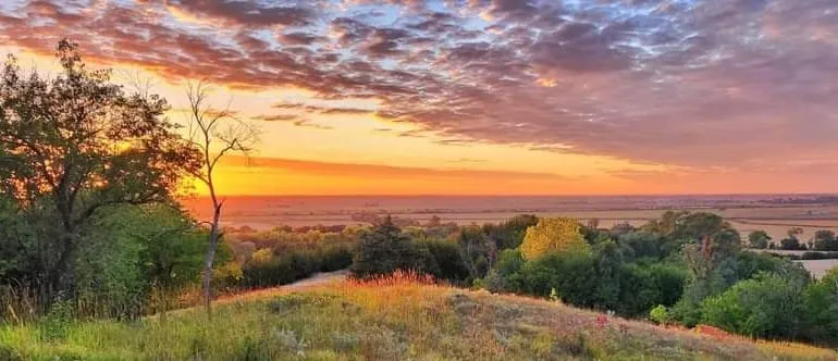 Sunset over a grassy hillside and wooded slopes, with open plains stretching to the horizon.