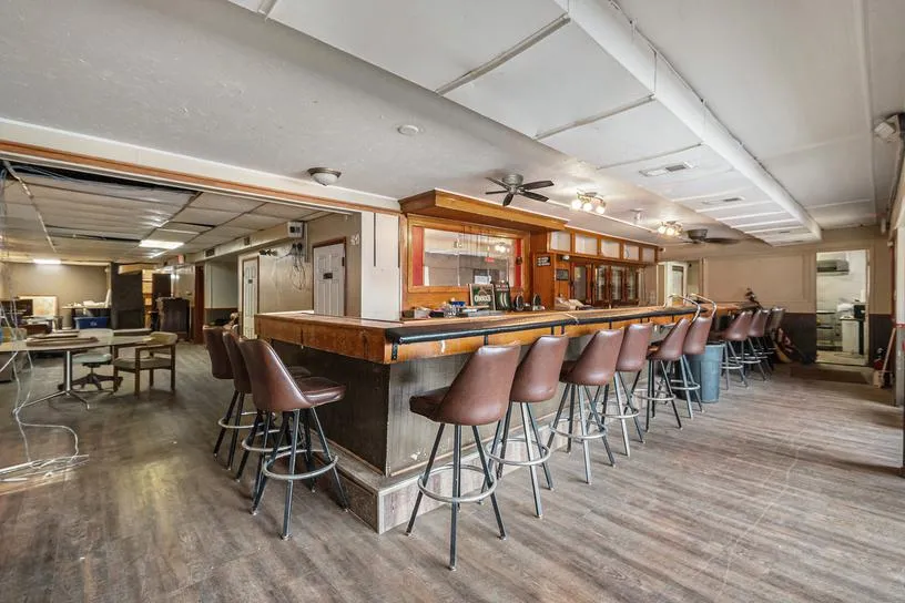 Bar interior with a long counter, rows of brown stools, and dining tables.