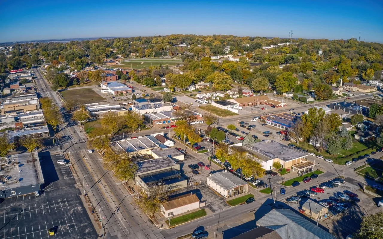 Aerial neighborhood view with low-rise shops, tree-lined streets, and a sports field.